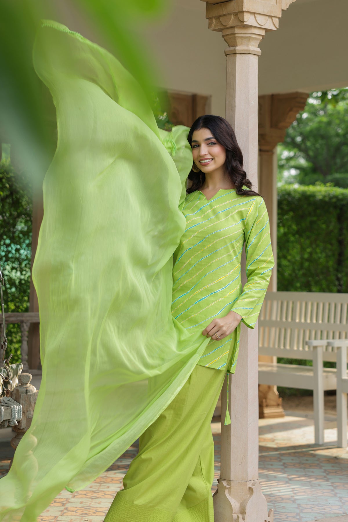 A woman modeling a bright lime green traditional outfit with a matching flowing sheer dupatta, standing in a garden pavilion setting.