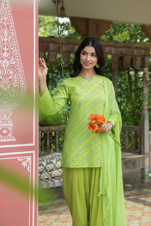 A woman modeling a bright lime green traditional three-piece salwar kameez suit with diagonal metallic stripes, standing in a garden pavilion setting and holding a small orange flower bouquet.