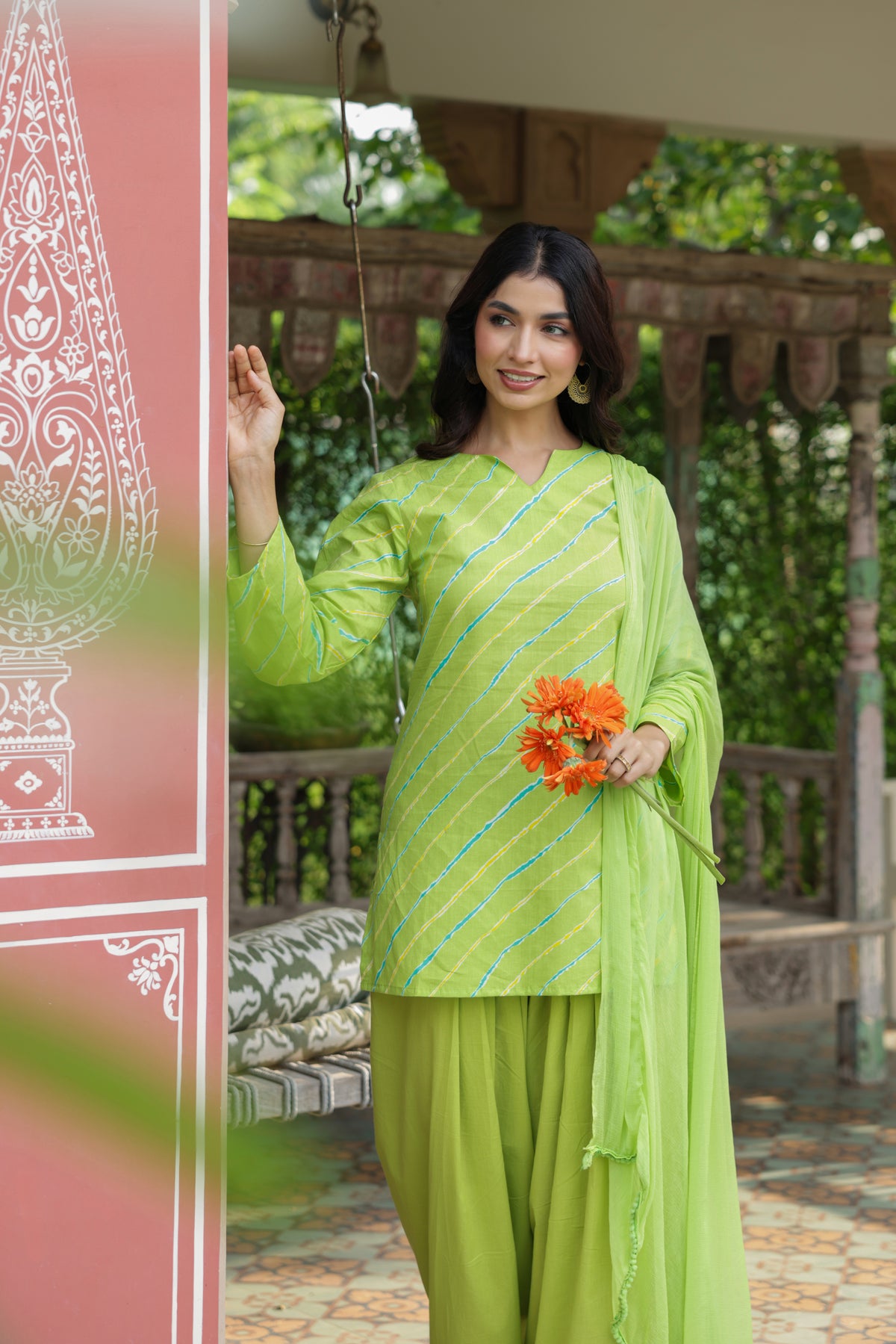 A woman modeling a bright lime green traditional three-piece salwar kameez suit with diagonal metallic stripes, standing in a garden pavilion setting and holding a small orange flower bouquet.