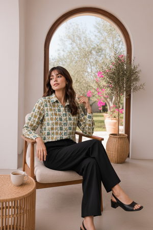 A woman sits in a wooden armchair wearing a long-sleeve button-up shirt with a multicolor geometric pattern in green, blue, and beige, paired with black trousers. A small side table with a coffee cup is in the foreground, and a potted plant is in the background.