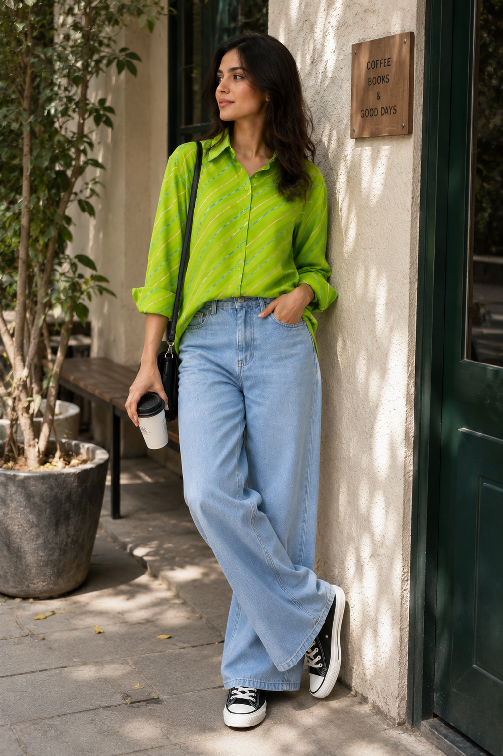 A woman stands on a sidewalk outside a shop, wearing a bright lime green striped button-up shirt, light-wash wide-leg jeans, black sneakers, and carrying a black shoulder bag while holding a takeaway coffee cup.