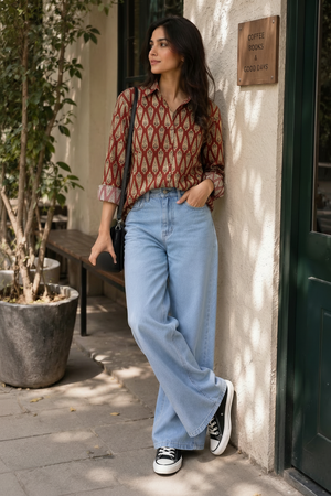 A woman stands outdoors against a beige wall next to a dark green door, wearing a red and brown patterned button-up shirt, light blue wide-leg jeans, black sneakers, and carrying a black shoulder bag while holding a white coffee cup.
