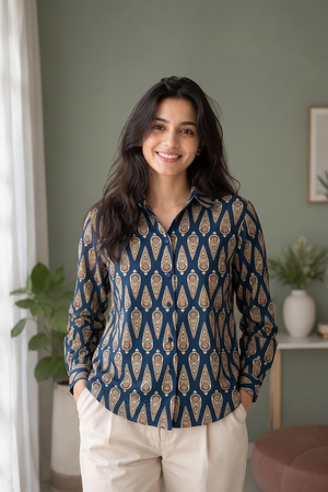 A woman wearing a navy blue long-sleeve button-up shirt with a beige and gold geometric pattern, paired with beige trousers, standing in a softly lit room with a green wall and indoor plants.