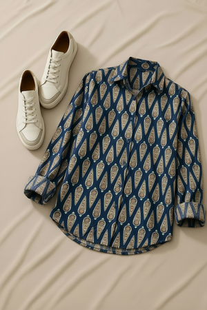 A navy blue long-sleeve button-up shirt with a beige and gold paisley pattern, styled on a black hanger next to a pair of white sneakers and a silver cuff bracelet on a beige fabric background.