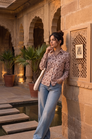 A woman stands in a stone courtyard wearing a long-sleeve button-up shirt with a pink and brown elephant print, light blue wide-leg jeans, and white sneakers, carrying a white shoulder bag.
