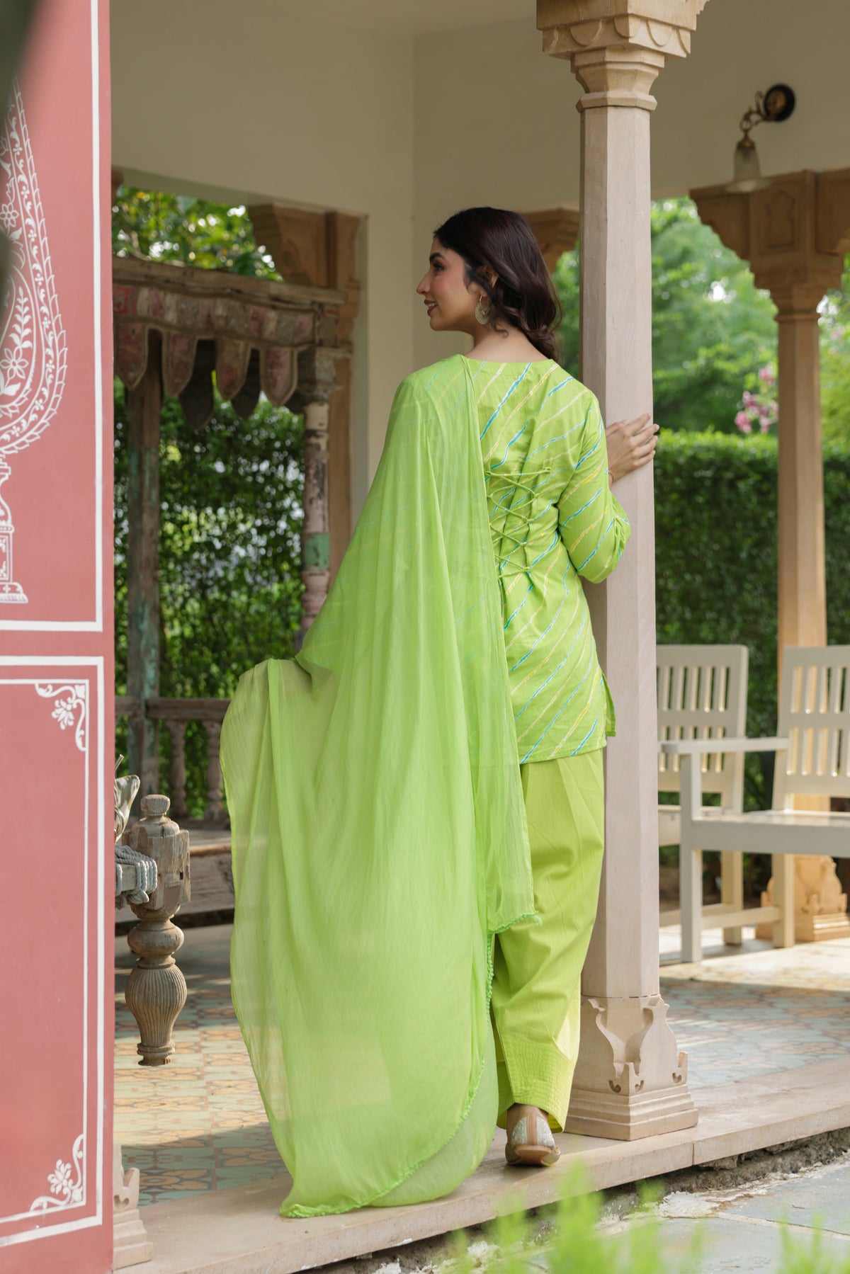 A woman wearing a bright lime green traditional outfit with a matching dupatta, standing on a covered porch with decorative pillars and greenery in the background.
