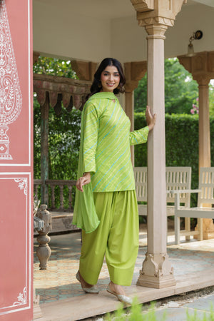A woman modeling a bright lime green traditional salwar kameez suit with diagonal striped patterns, standing in a traditional courtyard with beige pillars and green foliage in the background.