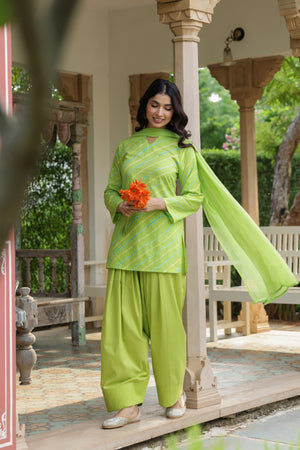 A woman models a bright lime green traditional three-piece outfit with matching dupatta, standing on a covered porch. The outfit features a kurta with diagonal stripes, wide-leg pants, and a flowing dupatta.