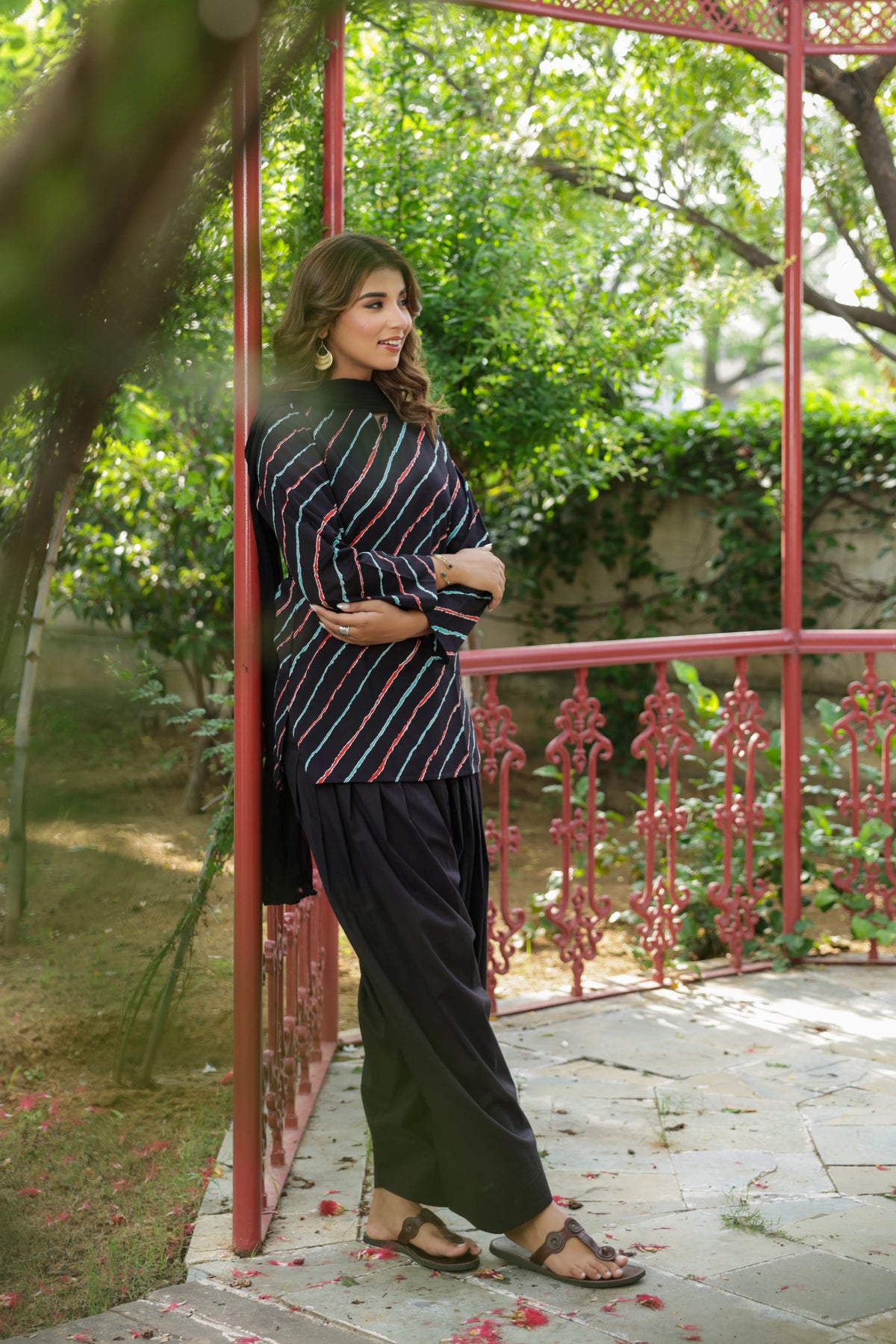 A woman stands outdoors in a garden-like setting beside a red metal trellis, wearing a dark striped long-sleeve top and wide-leg black pants with brown sandals.
