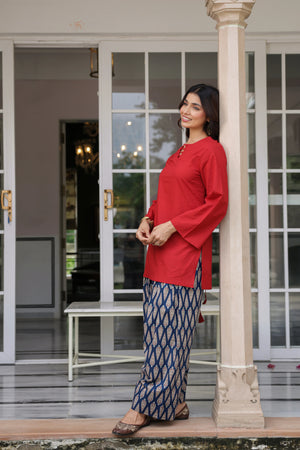 A woman wearing a bright red long-sleeve kurta and blue patterned wide-leg trousers stands leaning against a beige pillar in front of a white-framed glass door.