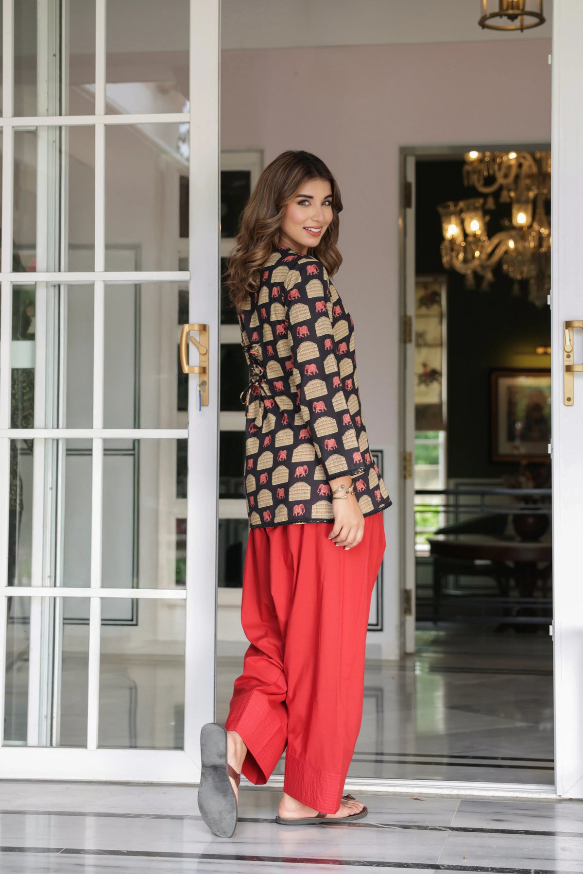 A woman wearing a patterned black kurta with red and beige motifs and bright red wide-leg pants, standing in a doorway of a modern home.
