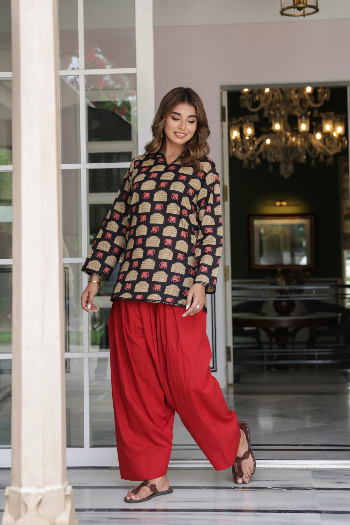 A woman modeling a traditional Indian outfit consisting of a beige kurta with a red and green pattern and wide-leg red pants, standing in a doorway of a house.