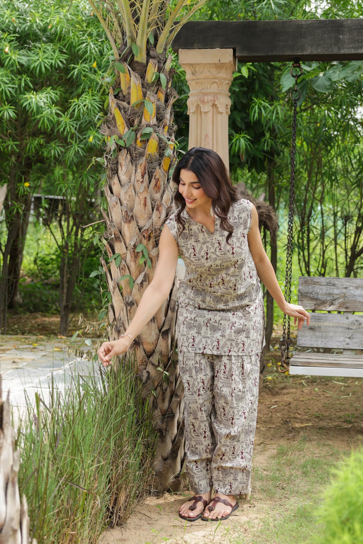 Aarika Beige Folk Print Cotton Farshi Salwar Set styled outdoors, featuring a sleeveless beige printed kurta with matching flowing Farshi salwar in a garden setting.
