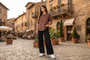 A woman stands on a cobblestone street in a European town, wearing a brown and black patterned button-up shirt, black wide-leg pants, and white sneakers.