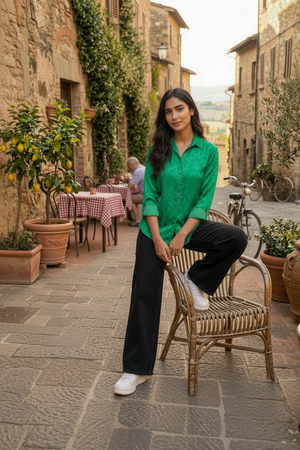 A woman posing on a rustic wicker chair in a sunlit, stone-walled outdoor restaurant alley. She is wearing a brown and black patterned button-down shirt, dark wide-leg jeans, and white sneakers.