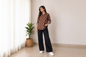A woman modeling a brown and black paisley print button-up shirt with wide-leg black pants and white sneakers, standing in a brightly lit indoor setting with a potted plant in the background.