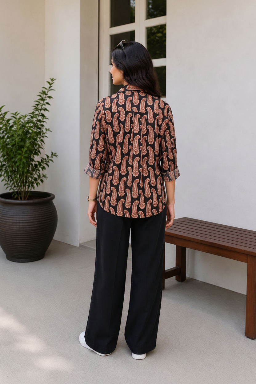 A woman stands with her back to the camera wearing a brown and black paisley patterned blouse with three-quarter sleeves and black wide-leg pants, set against a modern outdoor patio background with a potted plant and wooden bench.