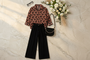A styled flat-lay outfit featuring a long-sleeve button-up shirt with a brown and red paisley pattern on a black background, paired with black wide-leg jeans, a black crescent shoulder bag, black low-top sneakers, and black eyeglasses, arranged on a light beige marble surface with white and yellow flowers.