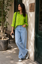 A woman stands on a sidewalk outside a shop, wearing a bright lime green striped button-up shirt, light-wash wide-leg jeans, black sneakers, and carrying a black shoulder bag while holding a takeaway coffee cup.