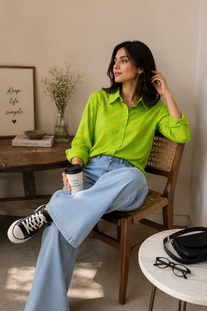A woman sits on a wooden chair in a bright room, wearing a neon green striped button-up shirt, light blue wide-leg jeans, and black-and-white sneakers. She holds a takeaway coffee cup, with a framed print, books, a vase, and a small table with glasses and a bag in the background.