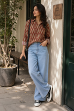 A woman stands outdoors against a beige wall next to a dark green door, wearing a red and brown patterned button-up shirt, light blue wide-leg jeans, black sneakers, and carrying a black shoulder bag while holding a white coffee cup.