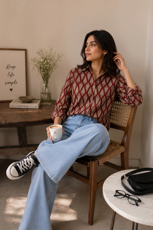 A woman sits on a wooden chair in a cozy room, wearing a red and gold patterned button-up shirt, light blue wide-leg jeans, and black Converse sneakers. She holds a takeaway coffee cup.