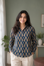 A woman wearing a navy blue long-sleeve button-up shirt with a beige and gold geometric pattern, paired with beige trousers, standing in a softly lit room with a green wall and indoor plants.
