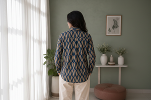 A woman viewed from behind wearing a long-sleeve button-up shirt with a navy blue and beige geometric pattern, paired with cream trousers, standing in a styled room with a green wall and plants.