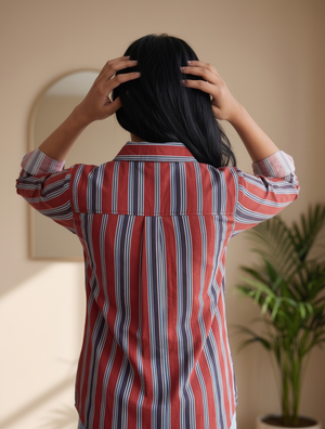 A person viewed from behind wearing a red, white, and blue vertically striped button-up shirt, with their hands in their dark hair, in a softly lit indoor setting with a potted plant in the background.