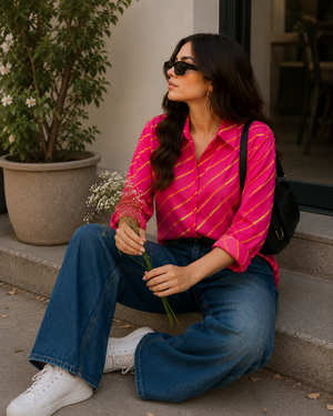 A woman sits on concrete steps wearing a bright pink striped button-up shirt, blue wide-leg jeans, white sneakers, and black sunglasses, with a black shoulder bag and holding small white flowers.