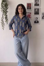 A woman stands indoors wearing a blue button-up shirt with a gold abstract pattern and light-wash wide-leg jeans. The background is a white wall with framed portraits and a hanging green plant.