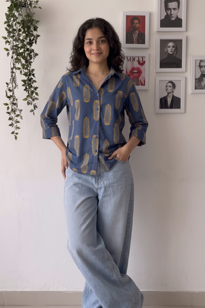 A woman stands indoors wearing a blue button-up shirt with a gold abstract pattern and light-wash wide-leg jeans. The background is a white wall with framed portraits and a hanging green plant.